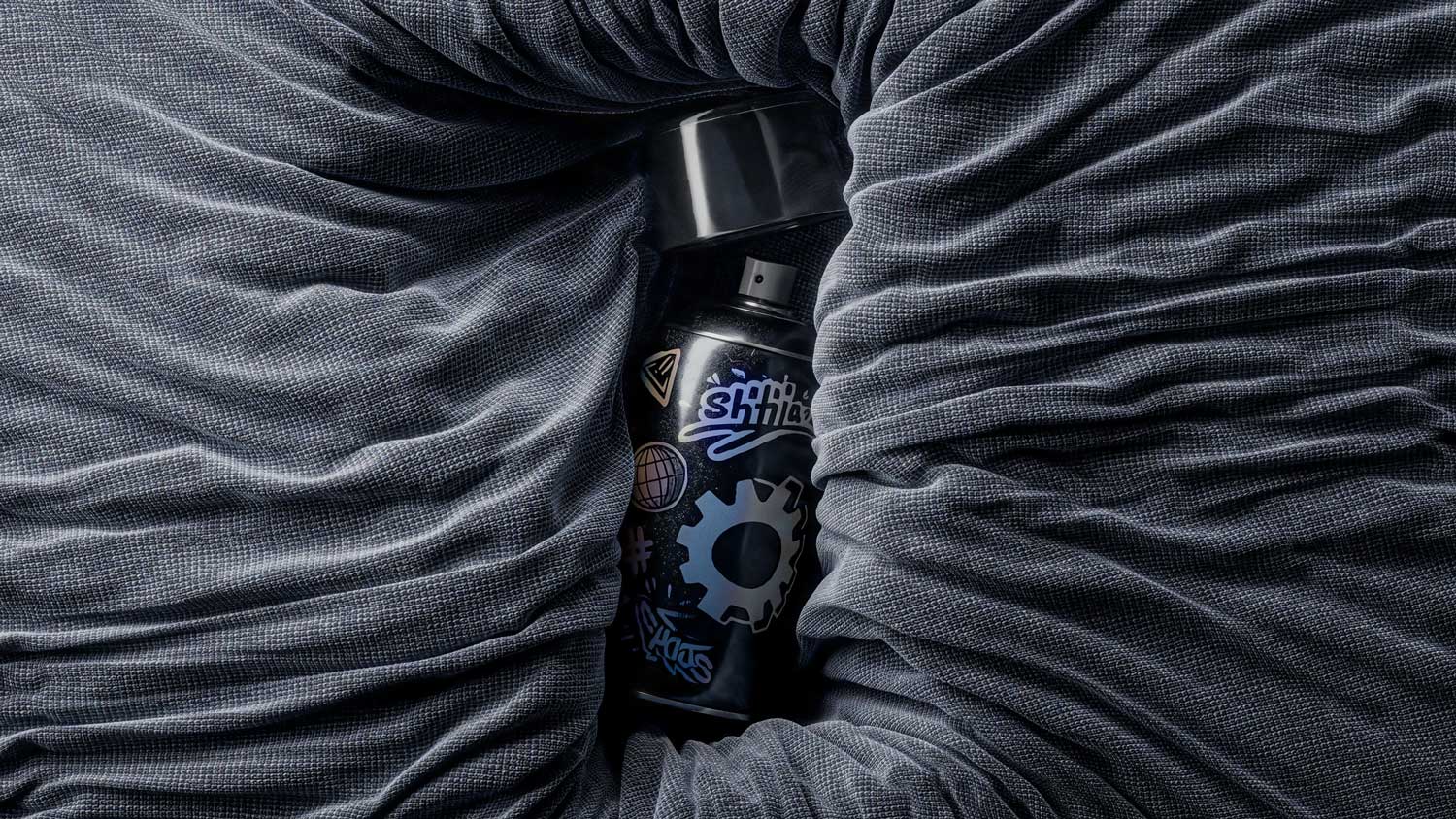 A CGI render of a black spray paint can with holographic stickers, including a gear and globe, nestled within dark grey, textured fabric. David Gobeev, rvre.graphics